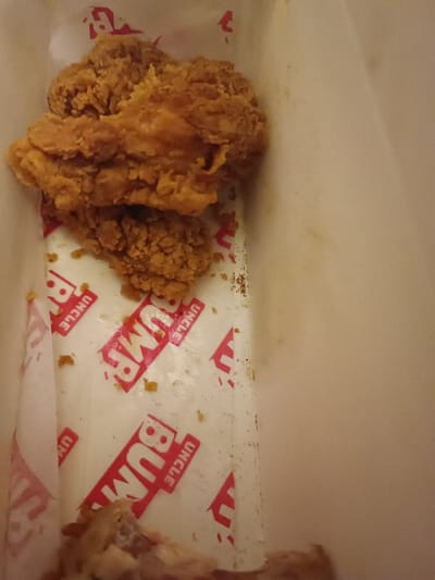 An opened white cardboard box contains leftover fried chicken. Three pieces, heavily breaded and golden-brown, sit atop white paper patterned with red lettering "UNCLE BUMP". Crumbs and small pieces of breading scatter the paper lining. At the bottom of the box, partially obscured, lie picked-clean chicken bones. The right side of the box is empty and coated with a thin oily residue.