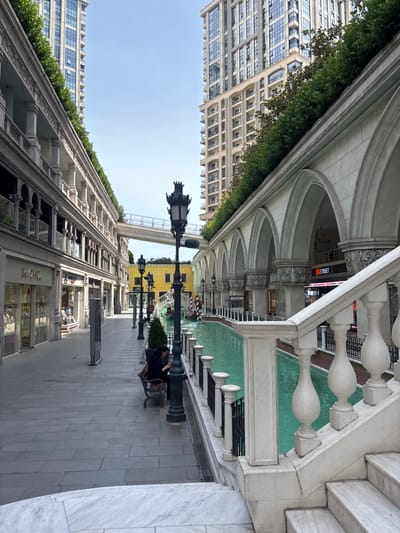 The image shows a canal running alongside a walkway lined with buildings designed in a Venetian architectural style. Tall, modern apartment buildings rise in the background. There are shops on the ground floor of the buildings lining the walkway, with signs such as "So CHIC" and "IN STREET" visible. A bridge spans the canal in the distance. A single person is seated on a bench on the walkway, looking at a mobile phone. The canal water is a vibrant turquoise color. The scene appears to be in a commercial or entertainment district. The location is the Venezia Mega Outlet shopping mall in Gaziosmanpaşa, Istanbul, Turkey, which is designed to resemble Venice.