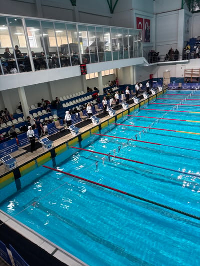The image shows an indoor swimming pool with several lanes separated by lane ropes, and flags hanging above the water. Each lane is marked by starting blocks with numbers. On the left side of the pool is a seating area filled with spectators. A two-story section with glass windows is visible, also occupied by people. A digital scoreboard is on the wall. A banner near the edge of the pool contains text that includes: "ISTANBUL GENCLIK," and "ISTANBUL GENCLIK VE SPOR." This suggests the location is in Istanbul, Turkey.
