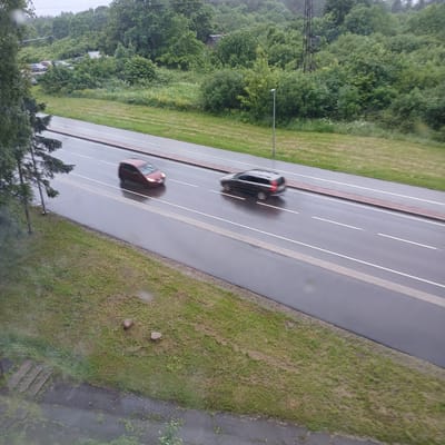 This is an aerial, slightly tilted, wide shot of a wet, paved road in Narva, Estonia, likely taken on a cloudy or rainy day. The road has white lane markings and a reddish-brown border on the left. Two cars are visible, both blurred due to motion. The car further up the road is dark, and the one closer to the viewer is a reddish-brown. The road is bordered on the left by a grassy verge with some trees and on the right by more grass and a paved area. In the background on the left, there are dense green trees and a tall, dark structure that appears to be a power pylon. Further in the distance, some parked cars are visible amongst the trees. The scene has a sense of everyday activity on a road, but the motion blur of the cars and the overcast lighting suggest a common, perhaps mundane, moment. There is no visible text in the image.