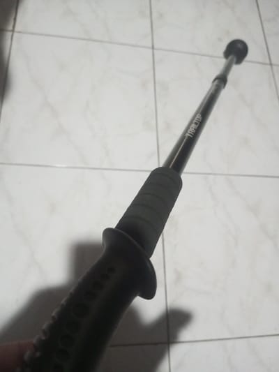 A black Trail Top trekking pole sits diagonally on light-colored, square-patterned tiles. The pole has a textured grip with finger grooves and a flange below the grip. The Trail Top logo is printed on the shaft. The top of the pole has a rounded rubber tip. A shadow is cast on the tiles.