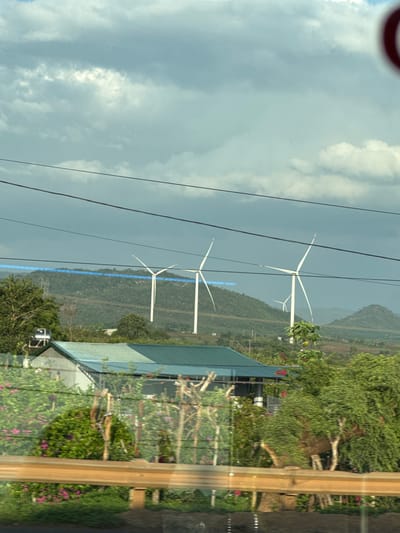 The image portrays a daytime landscape in Thôn 4, Vietnam, under an overcast sky. In the middle ground, three prominent white wind turbines with distinctive red-tipped blades are situated on a series of verdant, undulating mountains, indicating a wind farm. A striking blue line, possibly a road or a cleared path, runs horizontally across the distant mountain range.
Multiple power lines stretch across the upper part of the frame. To the lower right, a portion of a building with a dark green, angled roof and white walls is visible, nestled among lush green trees and bushes adorned with pink flowers. A water tank is also present near the building. The extreme foreground on the right displays a light brown wooden frame, likely part of a window or a vehicle's interior, showing reflections of the outside scene.
No specific human activity is depicted. The weather is cloudy but bright, suggesting mid-day. The scene captures a harmonious blend of natural beauty and modern renewable energy infrastructure in a rural, mountainous setting. No text is visible in the image.