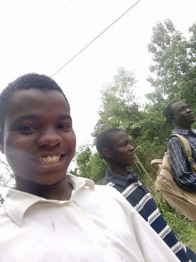 Three young men are depicted in an outdoor setting. The man in the foreground is smiling at the camera, wearing a white collared shirt. Behind him and to the right is another young man in a navy blue and white striped shirt. Further in the background is a third young man, wearing a blue plaid shirt and carrying a tan backpack. They appear to be standing near or on a road, with greenery and trees visible in the background. Based on the prompt, the location is assumed to be Eleweran, Nigeria.