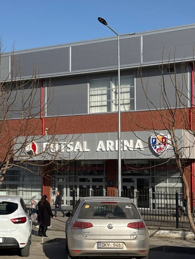 This image depicts the exterior of a "FUTSAL ARENA" building in Ciorescu, Moldova, under a clear blue sky. The multi-story structure features gray paneling on its upper levels and a red brick facade on the lower section. Prominently displayed above the main entrance are large white letters spelling "FUTSAL ARENA," flanked by a red and white futsal ball logo on the left and a colorful crest emblem on the right. The ground-level entrance consists of multiple glass double doors, with windows on the upper floor fitted with white vertical blinds. A red vertical pipe is visible on the building's left side.
In the foreground, a white SUV is partially visible on the far left, while a silver Volkswagen Golf hatchback (Moldovan license plate DKY 946) is parked centrally, facing away from the building. A tall, gray lamppost with an LED fixture stands in front of the building, slightly obscuring the facade. Three individuals are observable near the building's entrance: a person in a dark coat is walking towards the right, near the white SUV; another individual is partially visible behind and to the left of this person; and a man in a dark jacket is walking right, further into the frame, between the parked vehicles. Several deciduous trees with bare branches are present in the foreground and midground. The area in front of the building is paved, and a black metal fence borders the scene to the right. The lighting suggests a bright, clear day.