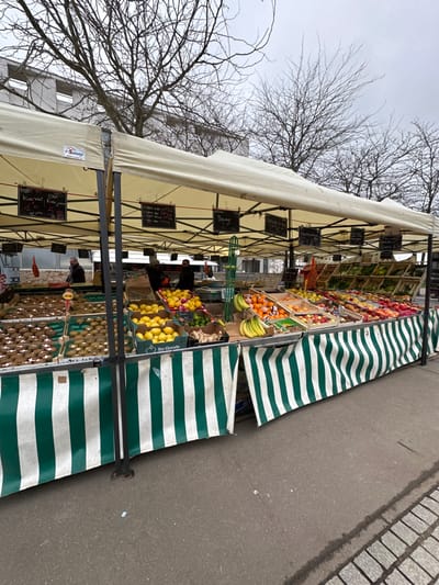 The image displays an outdoor fruit market stall operating in Serris, France. The stall features a cream-colored canvas canopy, with a visible "Sunny" brand logo on the left, supported by a dark metal frame. The front of the stall is adorned with a green and white vertically striped fabric skirting.

Various fruits are systematically arranged on the stall's surface and in wooden crates. Prominently displayed items include kiwis, lemons (some in crates marked "So Goods"), limes, bananas, oranges, grapefruits, and ginger roots. Additional fruits and vegetables are visible further along the display to the right. Black chalkboard signs with white handwritten text, suspended from the canopy, identify produce and list prices in Euros, such as "Kiwi Vert 1€50 les 500g," "Banane 2€ les 500g," "Orange à 2€ 1Kg," and "Pamplemousse à 1€80 Pièce."

Three individuals are partially visible behind the stall's counter, situated towards the left and center-left. Two are positioned near the fruit displays, while a third is further in the background to the left. The ground in the immediate foreground is dark gray asphalt, transitioning to a cobblestone surface on the far right. The background features several leafless trees, indicative of winter or early spring, positioned in front of light-colored, multi-story buildings, all under an overcast sky.