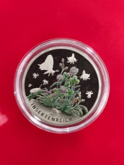 The image shows a close-up of a round, silver coin encased in clear plastic, resting on a bright red surface. The coin features a detailed relief of various insects and plants on a black background. Visible on the coin are a butterfly, a beetle, and several smaller flying insects, amidst depictions of clover flowers and blades of grass. The inscription "INSEKTENREICH" is visible along the lower edge of the coin. The red surface suggests a table or fabric. There is no discernible connection to Triberg im Schwarzwald, Germany, in the image itself.