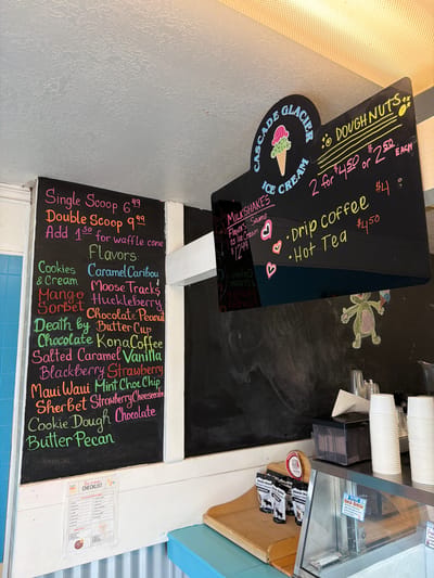 This media file captures a close-up view of a menu board and counter area in what appears to be an ice cream shop or cafe. The scene is dominated by two chalkboards displaying offerings, positioned against a light grey and white paneled wall.

The left chalkboard lists ice cream options and flavors. "Single Scoop" is priced at $6.49, and a "Double Scoop" at $9.99, with an additional $1.50 for a waffle cone. A diverse list of "Flavors" includes Cookies & Cream, Caramel Caribou, Mango, Moose Tracks, Sorbet Huckleberry, Chocolate Peanut, Death by Butter Cup, Chocolate KonaCoffee, Salted Caramel, Vanilla, Blackberry, Strawberry, Maui Waui, Mint Choc Chip, Sherbet Strawberry Cheesecake, Cookie Dough Chocolate, and Butter Pecan. A partially visible paper titled "Ice cream CHECKLIST" is also attached to the wall.

The right chalkboard, shaped like an ice cream cone, features the name "CASCADE GLACIER ICE CREAM." It advertises "MILKSHAKES" for $12.49, noting that the "Flavors" are "Same as Ice Cream." "DOUGHNUTS" are offered at "2 for $4.50" or "$2.50 EACH." Beverages include "Drip coffee" for $4.00 and "Hot Tea" for $4.50. A whimsical drawing of a green frog holding an ice cream cone is visible on the wall behind this chalkboard.

On a counter to the right, stacks of white disposable cups are arranged next to a clear display case. Inside the case, a small sign for "Birch Brittle" describes it as "with 100% pure late run birch syrup & pieces of birch brittle." Small packaged snacks, possibly "Moose Bites," are also visible in a wooden holder. The setting suggests a casual, local establishment focused on frozen treats and simple beverages.