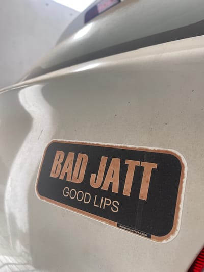A close-up, low-angle shot captures a sticker on the trunk of a light-colored vehicle, likely in Sangrur, India. The sticker is rectangular with rounded corners and a distressed brown border. The top line of text on the sticker reads "BAD JATT" in large, blocky, distressed brown letters. Below this, in smaller, distressed brown sans-serif font, is the text "GOOD LIPS". In the bottom right corner of the sticker, a small black bar contains white text that reads "WWW.URBANTHEKA.COM". The vehicle's surface appears somewhat dusty. The background is blurred, showing parts of the car's trunk and a hint of a taillight on the right. The overall lighting suggests an outdoor setting, possibly during the day.