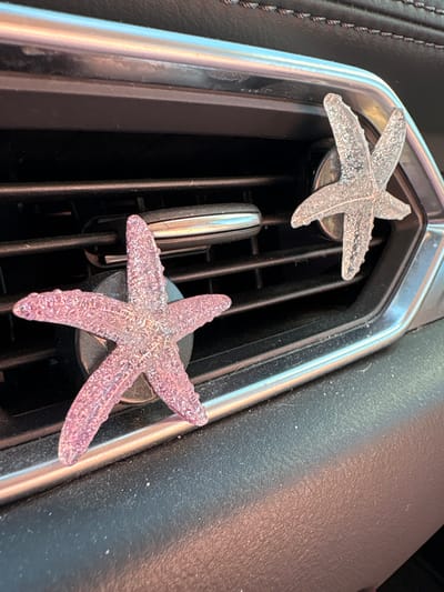 The image shows the dashboard air vent of a car. Two decorative starfish are clipped to the vent. The upper starfish is translucent and colorless. The lower starfish is translucent and light pink. The car’s dashboard is visible on the right side of the image, and a stitched leather section can be seen on the left. This is a generic scene that could be found in any car, and no definitive indicators place this scene specifically in Huntington Beach, California.