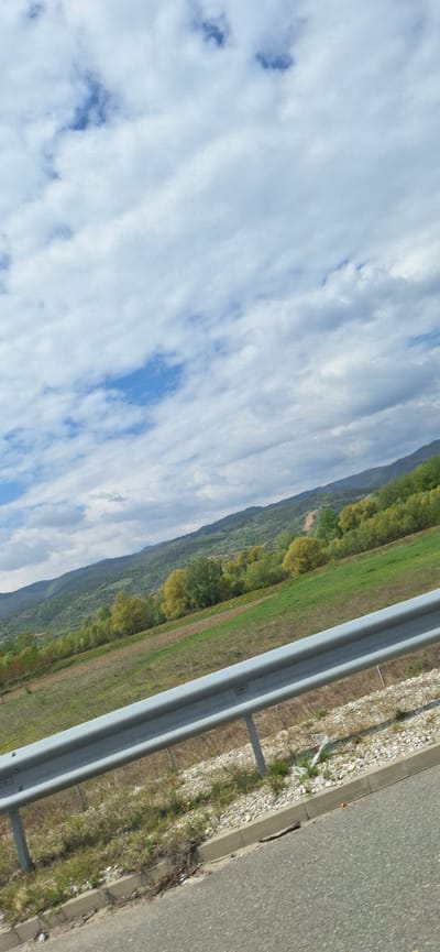 The image captures a scene along a highway near Valkovo, Bulgaria. In the foreground, a gray asphalt road is visible with a concrete curb and gravel shoulder. A silver metal guardrail runs parallel to the road, separating it from a green field. Beyond the field, a wooded hillside rises, its trees showing variations in color. The sky is filled with white clouds, interspersed with patches of blue. There are no visible people or actions in the scene. The focus is on the landscape and infrastructure.