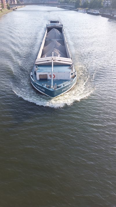 This is an aerial, top-down drone shot of a cargo ship named "STAR FLYER" navigating a wide, dark river, likely the Meuse River in Namur, Belgium. The ship is moving from left to right across the frame, creating a significant wake behind its bow, indicative of a moderate speed. The ship is laden with a cargo of light-colored aggregate, possibly sand or gravel, filling its open hold.

The overall scene suggests a daytime setting, with sunlight reflecting off the water and the ship's surfaces, creating bright highlights and casting subtle shadows. The weather appears calm and clear, with the water exhibiting small ripples rather than strong waves. The left side of the frame shows a glimpse of a riverbank with some buildings and trees, suggesting an urban or semi-urban environment. The right side of the frame is dominated by the dark, rippling water.

The text "STAR FLYER" is clearly visible in white lettering on the port side of the ship's bow. There is also a red life preserver visible on the deck. The image captures the functional aspect of river transport, with the cargo vessel being the central focus as it efficiently moves its load through the waterway. No people are clearly visible, but their presence is implied by the operation of the ship. The overall mood is one of industriousness and transit.