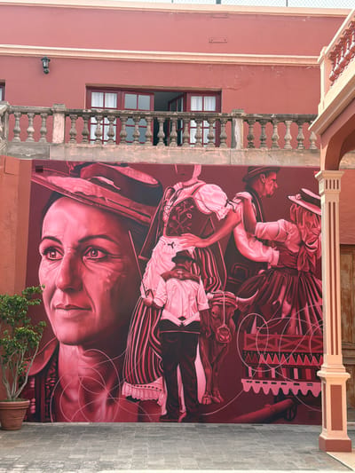 This is a photograph of a large, monochromatic mural painted in shades of red and pink on the side of a building in La Orotava, Spain. The mural depicts a scene with several figures dressed in traditional clothing and a bull. On the left side of the mural, a large portrait of a woman's face is visible, looking to the right. To her right, several other figures are depicted in motion. One figure, a man from behind, is dressed in a white shirt and black pants, and appears to be standing near the bull. Two other figures, a man and a woman, are dancing or interacting closely, both wearing hats and traditional garments. A bull is depicted in the center of the mural, facing left. In the background of the mural, elements of traditional carts or structures are visible. The building behind the mural has a reddish-pink facade, with a stone balustrade and windows on an upper level. A small potted tree with green leaves is situated to the left of the mural. The ground in front of the mural is paved with square stone tiles. The overall scene suggests a representation of local culture or historical events.