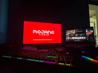 The image depicts a dimly lit indoor computer setup located within a residence or personal space in Straldzha, Bulgaria. The scene is primarily illuminated by the glow of electronic displays and multicolored RGB peripherals.
In the center, a large monitor displays a bright red screen with the white text "MOJANG" prominently featured above "STUDIOS." Below this, a white loading bar is visible, approximately three-quarters full, indicating the initialization sequence of a game or application from Mojang Studios, likely Minecraft.
To the right of the main monitor, a laptop is open and powered on, functioning as a secondary display. Its screen shows a dark-themed application, likely a media player or game launcher. Visible content includes multiple small square thumbnails of various images, some appearing to be album art or game icons. Partially discernible Cyrillic text reads "Всичко ново, което трябва да чуеш!" (Everything new you need to hear!), suggesting music recommendations, and "DISCOVER WEEKLY" is also visible. The laptop's integrated keyboard features red backlighting.
On the desk in front of the monitors, at least two distinct keyboards are present. The left-most keyboard is full-sized, black, and features active RGB backlighting with colors transitioning from green to red, purple, and blue across its keys. A second keyboard section, or an extension of the first, is positioned to its right, also displaying RGB lighting in various hues, including green, blue, pink, and yellow. A computer mouse, equipped with vibrant RGB lighting that creates a rainbow trail effect, is positioned to the right, below the laptop.
A translucent, possibly plastic or fabric, cover is draped over the base of the main monitor and extends slightly over the rear of the keyboards. Cables are visible connecting the peripherals. The wall to the left appears dark with a subtle textured pattern, while the wall to the right is lighter in color, showing a corner and some faint discoloration or wear near the top-right. No individuals are visible in the scene.