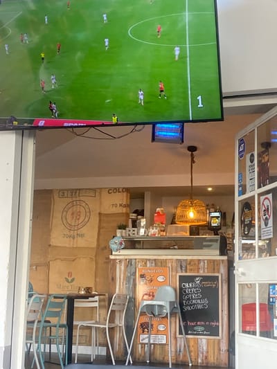 The image depicts the interior of a cafe, possibly in San Pedro De Alcántara, Spain, with a television screen displaying a football match in the background. The cafe features a rustic wooden counter and walls covered with large sacks, some marked with "Mare Terra" and "70 Kg Net". A blackboard menu lists items such as "CHURROS", "CREPES", "GOFRES", "BOCADILLOS", and "SMOOTHIES". There are tables and chairs, including a high chair for children. The overall ambiance suggests a casual dining establishment.