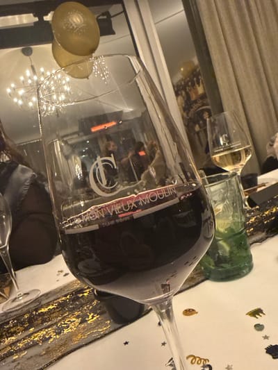 This media file captures an indoor scene at what appears to be a celebratory event in Grass, Luxembourg, likely in the evening or night. The primary subject is a very large wine glass, filled with red wine, angled across the frame. Reflections within the glass show a modern chandelier and glimpses of a person. To the left, another, smaller wine glass likely holds white wine. A green glass bottle is also visible on the table.
The table surface is adorned with a white cloth, scattered with festive confetti in gold and black, including star and spiral shapes, suggesting a party or special occasion. Another golden, circular decorative object is partially visible on the left.
Visible text on the large wine glass includes "DON VIEUX MOULIN" and "DUHR frères," indicating a specific wine or brand. A stylized logo, possibly a "D" or "C," is also present. The scene implies a social gathering with drinks, set in a festive, well-lit environment.