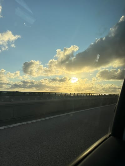 The image depicts a roadside view with a sunset in the background. The foreground shows a grey asphalt road with a white line marking the lane. To the left, a concrete barrier with a black metal railing runs along the edge of the road. Beyond the barrier, a distant landscape with trees and some structures is visible. The sky is filled with a mixture of blue sky and fluffy, golden-lit clouds, with the sun setting behind a large cloud formation. Reflections from a window are visible in the upper part of the image, obscuring some of the sky. The scene appears to be a highway or an elevated road, possibly in or near Ödsmål, Sweden, given the presence of wind turbines in the distance. There are no people visible in the image.