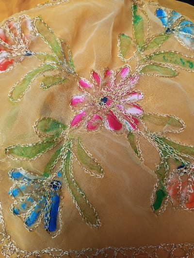 The image showcases a piece of golden-yellow fabric adorned with colorful floral patterns. Flowers in shades of pink, blue, and orange are scattered across the material, each outlined with delicate gold embroidery. Green leaf-like shapes accompany the flowers, also defined by the same gold thread. The fabric is light and slightly sheer, lending a soft and elegant look to the overall design.