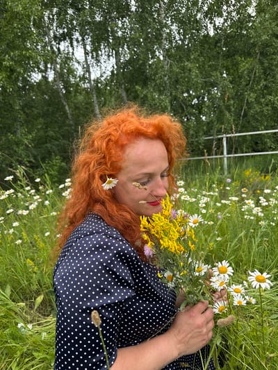 A woman with bright red, curly hair is lying down in a grassy field with wildflowers. She is wearing a dark blue dress with white polka dots. Her eyes are closed, and she appears to be relaxed, with a slight smile on her face. She is holding a bouquet of yellow wildflowers and daisies. A single daisy is tucked into her hair above her ear.

The scene is outdoors, with a background of green trees. The lighting suggests it is daytime, likely a sunny day given the vibrant colors of the flowers. The overall mood is peaceful and serene. There are no visible text elements in the image. The location context is Privolzhsky, Russia.