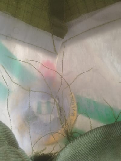 This is a close-up, abstract image of layered fabrics, with no discernible landmarks or elements that would indicate a specific location in Majemu, Nigeria. The image features a textured green fabric in the foreground and background, with a white, translucent layer in between. Thin, wavy green threads are scattered across the white layer, some appearing to be stitches or loose threads. A blurred, pinkish-red shape is visible beneath the white layer, along with streaks of green and yellow. The overall impression is one of textile construction or detail, devoid of any context related to a city or its inhabitants.