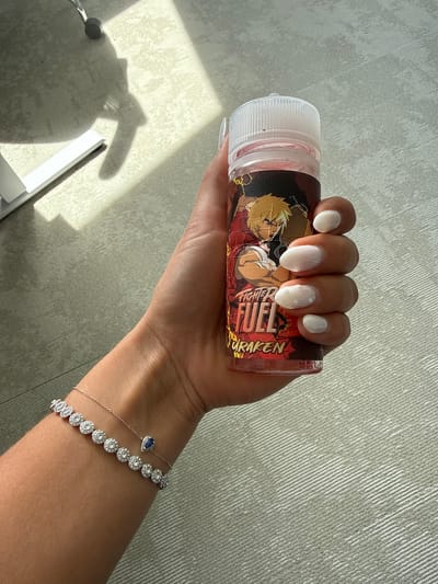 A person is holding a bottle of "Fighter Fuel Furaken" e-liquid in their right hand. They are wearing a silver bracelet with several round white gems and a separate silver chain bracelet with a dark blue teardrop gem. The nails on their hand are painted a glossy, off-white color. The bottle features an anime-style character and the words "FIGHTER FUEL FURAKEN." The background appears to be a textured gray surface. The location of Les Arcs, France cannot be confirmed based on the visual information provided.