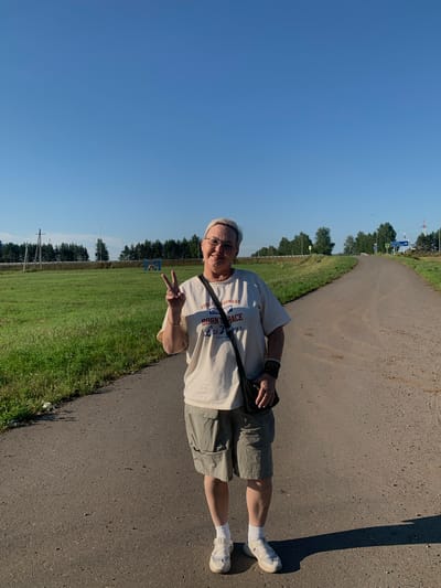 A person stands on an asphalt road in an outdoor environment, possibly in the city of Sokolka, Russia, on a clear day. The person, who appears to be female, is wearing a light-colored t-shirt with text and graphics, grey shorts, white socks, and white sneakers. They are also wearing glasses and a black crossbody bag. The person is smiling and making a peace sign with their right hand. The road stretches out ahead of them and curves to the right. To the left of the road is a grassy field, and in the distance, there are trees and power lines. The sky is a clear, bright blue. There are some road signs visible in the distance, but they are too small to read clearly. The overall scene suggests a casual outdoor setting.