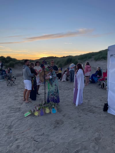 A group of people are gathered on a sandy beach at dusk. The sky is a pale blue with streaks of orange and pink from the setting sun. In the background, sand dunes covered in tall, green grass are visible. A white tent is set up on the right side of the frame.

In the foreground, several people are standing around a central figure dressed in a long, patterned dress and a flower crown. This person appears to be holding and playing a large, round drum decorated with colorful ribbons. Other individuals are observing the activity, some standing, some sitting in folding chairs. One man in a grey hoodie and white shorts is standing near the drummer, with his arm around a woman. Another woman in a white turtleneck and a person in a purple dress with a white cape are also near the center. Several small, decorated paper bags are placed on the sand in front of the drumming area. The overall atmosphere seems peaceful and ceremonial.
