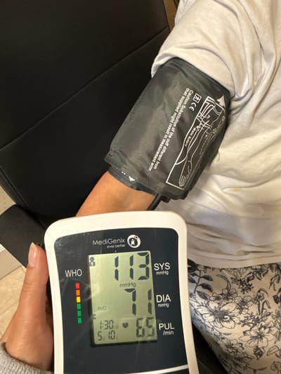 An indoor, eye-level shot captures a person taking their blood pressure. The subject is wearing a light-colored long-sleeved top and patterned trousers with a floral design, seated in a black chair. A grey blood pressure cuff is wrapped around their upper arm. Visible on the cuff are diagrams for "Main Artery" placement, a "CE" mark, and a caution text reading: "Caution: Substitution of the cuff different from that supplied might result in measurement error."
The person's hand, with neatly manicured nails, holds a white and dark grey "MediGenix ever better" blood pressure monitor. The monitor's display shows the current readings: Systolic (SYS) at 113 mmHg, Diastolic (DIA) at 71 mmHg, and Pulse (PUL) at 65 beats per minute. A timestamp of "1:30 5/10" is also visible. Below the readings, a "WHO" (World Health Organization) indicator bar displays green segments, suggesting the blood pressure falls within a healthy range. The overall scene indicates a routine health monitoring activity.