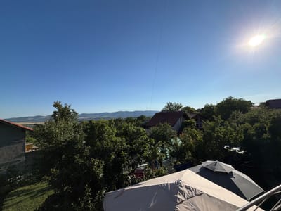 This is a high-angle, wide-shot photograph taken from an elevated position, likely a balcony or a higher floor of a building, overlooking a rural or semi-rural residential area in Golemo Buchino, Bulgaria. The scene is bathed in bright sunlight, with the sun positioned in the upper right corner of the frame, creating a lens flare and casting distinct shadows.
The foreground is dominated by two large outdoor umbrellas. The closest umbrella, in a light beige color, occupies the lower right portion of the image. Partially overlapping it is a darker gray and black umbrella. A metal railing is visible to the far right, suggesting the viewpoint is from a structure with a balcony or deck.
Behind the umbrellas, a dense canopy of green trees fills the midground. Interspersed within the foliage are glimpses of residential buildings with red-tiled roofs. One building on the left, partially visible, has a dark gray, textured facade and a red-tiled roof. Further into the scene, the landscape transitions into rolling hills and mountains in the distance, under a clear, deep blue sky. A thin, dark wire runs diagonally across the sky from the top center towards the right.
The overall impression is one of a sunny day in a village setting, with a focus on outdoor living spaces and the natural surroundings. There are no people or animals clearly visible in the image. The image captures a serene, daytime view of Golemo Buchino.
