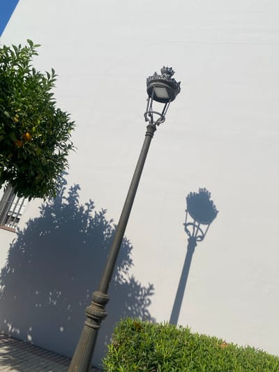 An ornate, dark metal street lamp stands at a diagonal, leaning slightly to the left, against a stark white wall in Cancelada, Spain. The lamp features a crown-like decorative top and a clear glass enclosure for its light. To the left of the lamp, a portion of a lush green tree with visible oranges is visible, casting a dappled shadow on the white wall. In the foreground, a neatly trimmed green hedge runs along the bottom edge of the image. The ground beneath the hedge appears to be paved with light-colored square tiles. A distinct shadow of the street lamp is cast on the white wall to the right of the actual lamp, and the shadow of the tree is cast on the wall to the left. The sky, a bright blue, is visible in the upper left corner of the frame, above the white wall. The scene is brightly lit by sunlight, creating strong contrasts between light and shadow.