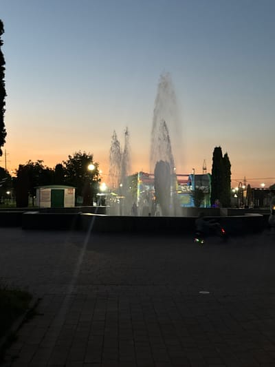 The image depicts a park scene at dusk, with a large fountain as the central element. The sky displays a gradient of colors, transitioning from a light blue at the top to warm orange and yellow hues near the horizon. Several water jets from the fountain are visible, creating a misty effect. In the background, there are trees, a small building with a green door, and what appears to be an amusement ride with colorful lighting. The ground in the foreground is paved with cobblestones. A person is seen on a wheeled device with illuminated lights, moving across the paved area. The overall atmosphere suggests a recreational space in the city of Endirey, Russia, during the evening hours.