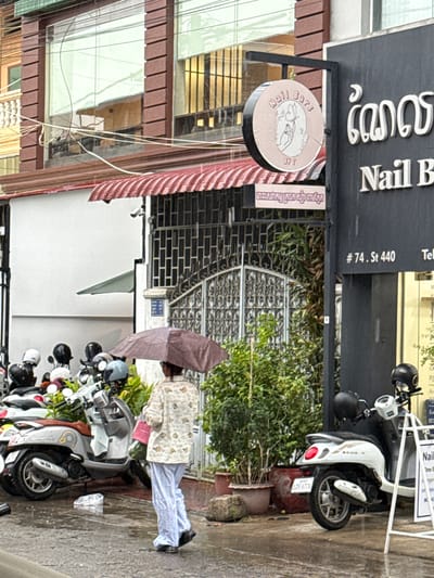 This is a photograph taken in Khan Chamkar Mon, Cambodia. The scene depicts a street in front of a business, likely a nail salon, indicated by the signage with text in both English and Khmer. The business name appears to be "Nail B" or similar, and the circular sign includes the words "Nail Bars" and a graphic of hands. There is also a sign with the address "#74. St 440".

Several scooters are parked along the sidewalk, some with helmets on them. A person is walking under a large, light-colored umbrella, suggesting it is raining or the sun is very strong. The person is wearing a patterned jacket and light blue pants. The building facade is visible, with glass windows and architectural details. The overall atmosphere suggests a typical urban street scene in Cambodia, possibly during the day, with overcast or rainy weather. The presence of parked vehicles and shop signage points to a commercial area. The text on the signs provides specific details about the business and its location.