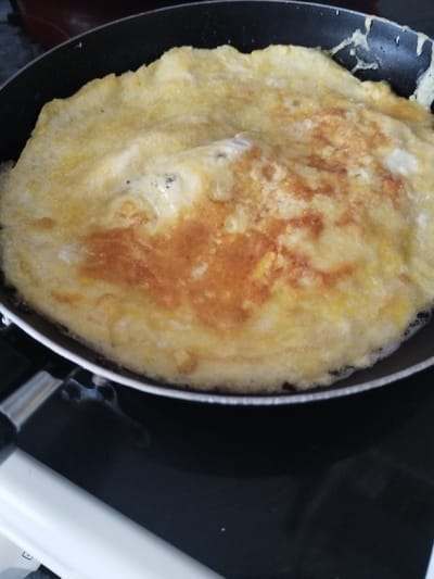 A close-up, overhead shot shows a golden-brown omelet cooking in a black frying pan. The omelet is slightly browned in some areas, indicating it is well-cooked. The pan is on a black stovetop, with a white appliance visible in the lower left corner. The background is out of focus and shows a hint of a red appliance. The image was taken in Novostroika, Russia.