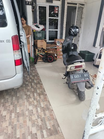 A silver minivan with its door open is parked beside a tiled area. A woman in a brown shirt stands near the minivan. Behind the minivan is a bicycle, a stack of cardboard boxes, and a basket holding white items. A black and gray moped is parked on the right side of the image. It has a helmet on the seat. A green plastic basket is sitting near the door. A white railing is in the lower right corner.