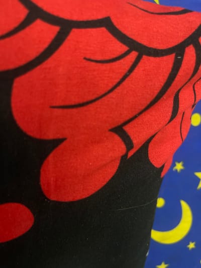 This image is a close-up, top-down shot of a fabric with a patterned design. The fabric is red with thick black outlines, forming a large, abstract floral or organic shape. The red is a vibrant, almost scarlet hue. To the right of the red patterned fabric, there is a section of black fabric. In the background, a blue fabric is visible, decorated with yellow crescent moons and stars, suggesting a celestial or nighttime theme.
The fabric appears to be slightly rumpled, creating folds and shadows that add depth to the image. The texture of the fabric is visible, with a slight weave discernible. There are a few stray, fine hairs visible on the black fabric. The lighting suggests it's an indoor setting, possibly a bed or a draped piece of material. No people are visible, and the focus is entirely on the textile design and its textures. The overall impression is one of a decorative fabric, perhaps from bedding or clothing, with a playful and whimsical pattern in the background. The provided location context of Anggaswangi, Indonesia, does not directly influence the visual description of the image itself, as no specific landmarks or cultural elements are identifiable within the frame.