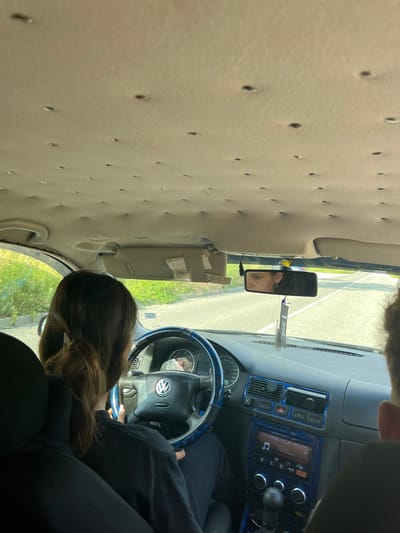 The image depicts the interior of a car, likely a Volkswagen Golf IV, as seen from the passenger seat. A young woman with brown hair tied in a ponytail is driving. She is wearing a black t-shirt and dark pants. Her hands are on the steering wheel, which has a blue and black patterned cover. The car's interior features blue accents on the dashboard and around the radio. The car is in motion, indicated by the road visible through the windshield and the reflection in the rearview mirror. The rearview mirror shows the face of a young woman with dark hair and earrings. The ceiling of the car has numerous small holes. The car appears to be driving on a paved road in an outdoor environment, with greenery visible on the left side of the frame. The location is identified as Taran, Bulgaria, based on the prompt.