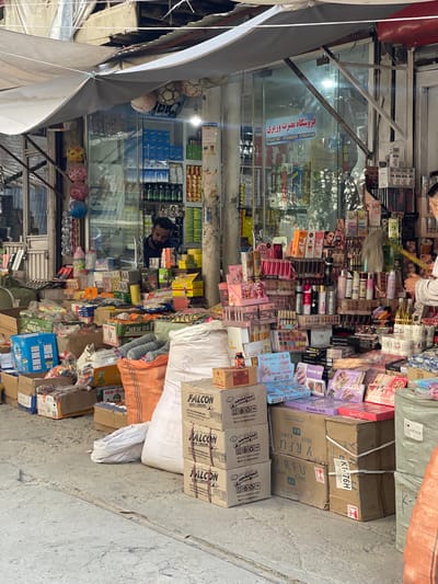 The image shows a bustling street market in Kabul, Afghanistan. The scene is dominated by a small shop with a glass storefront, packed with an array of goods. Various items are displayed on shelves inside and piled high in front of the shop, creating a sense of abundance and commercial activity.

Among the merchandise are cosmetics, skincare products, perfumes, and possibly household items. The storefront itself is adorned with signage in what appears to be Arabic script, advertising the shop's wares. The shopkeeper, a man with a beard, is visible behind the counter, engaged with customers or organizing inventory.

In front of the shop, an assortment of boxes, bags, and containers are stacked on the ground, indicating the sale of a diverse range of products. Some of these appear to be consumer goods, such as shoe cream and various packaged items, while others are less identifiable. The ground is paved with concrete, typical of urban street environments.

The overall impression is one of a vibrant marketplace where commerce is actively taking place. The market is outdoors, with a canopy providing some shade over the stalls and merchandise. The presence of everyday goods suggests this is a local retail area catering to the needs of the community in Kabul.