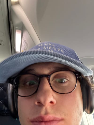 A person wearing a blue beanie, black-rimmed glasses, and headphones is facing the camera in a close-up shot. The individual's face is partially visible, showing one eye, nose, and mouth. The background includes an airplane cabin interior with ceiling panels and a reading light. The photo is taken at an angle.