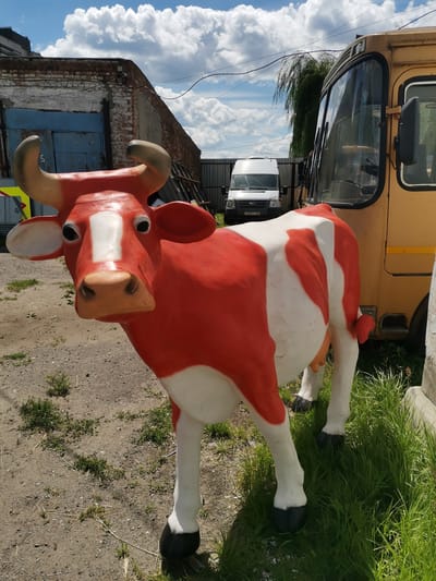 A red and white cow statue stands in an outdoor industrial area. The statue is life-sized and appears to be made of fiberglass or plastic. It has black hooves, prominent horns, and a realistic facial expression. The background features a yellow bus, a white van, a brick building with blue doors, a metal fence, and a cloudy blue sky. The ground is covered with gravel and sparse vegetation. This scene is located in the city of Rasskazovo, Russia, likely an industrial or agricultural facility. There are no people visible in the image, and no actions are taking place.