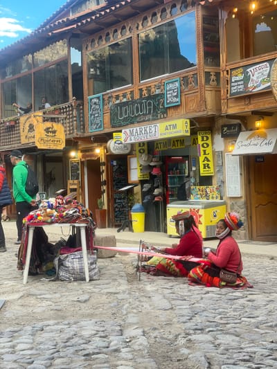 The image depicts a street scene in Ollantaytambo, Peru. Two indigenous women are weaving fabric on the sidewalk, positioned near a shop with signs indicating money exchange (ATM, EURO, DOLAR). Colorful hats and textiles are displayed on a table in front of them. The storefront of "Uchucuta Restaurant Market & Souvenir" is visible with multiple signs, including a "Veggie Healthy Delicious Menu" sign. Another restaurant, "Mijunapaq," is also indicated by a wooden sign. The building architecture is a mix of wood and stone. A person wearing a green shirt and carrying a black backpack stands near the textile display. The street is paved with rough stones.