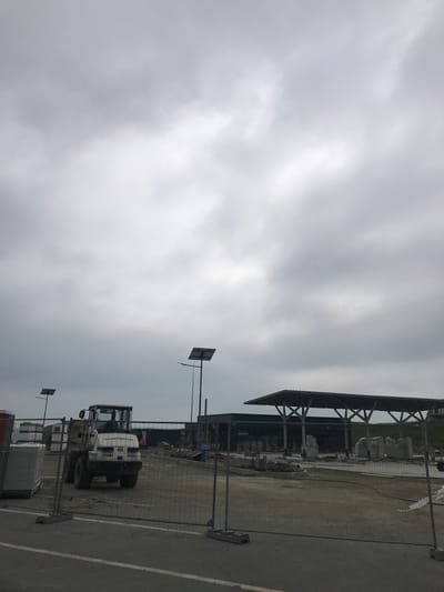 The image depicts an undeveloped or under-construction site in Șcheia, Romania, under an overcast sky. In the immediate foreground, a segment of an asphalt road with a single solid white line is visible. Behind this, a temporary chain-link fence, supported by concrete bases, spans the width of the frame.

Beyond the fence, a white wheel loader, branded "CATERPILLAR" on its cab and "www.AS-TRUCKS.de" along with the number "047" on its rear, is parked on a dirt and gravel lot. To the right of the loader, a large, dark, single-story building with a distinctive, wide overhanging canopy is present. The canopy is supported by several prominent X-shaped structural beams. Beneath and around this canopy, various construction materials are staged, including several large, rough-hewn concrete or stone blocks and stacks of wrapped pallets. A blue sign with white text is affixed to the temporary fence near the building.

Two solar-powered streetlights are visible; one in the mid-left, extending above the construction vehicle, and another in the mid-right, positioned near the building. The background consists of a gently sloped, earthen bank to the right and a uniformly grey, heavily clouded sky occupying approximately the upper two-thirds of the image. No individuals are present in the scene.