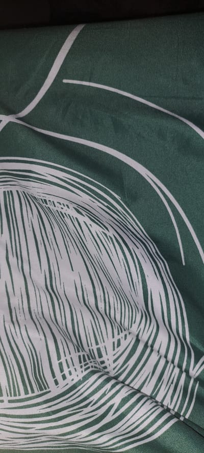 This is a close-up, abstract shot of a green fabric with a white, stylized palm leaf pattern. The fabric is folded and creased, creating shadows and highlights that emphasize the texture and the curves of the design. The palm leaf motif is rendered in bold white lines against the deep green background, with some of the lines appearing thin and delicate, while others are thicker and more solid. The image has a soft focus, adding to the artistic and textured feel. There is no discernible activity, time of day, weather, location cues, or visible text.