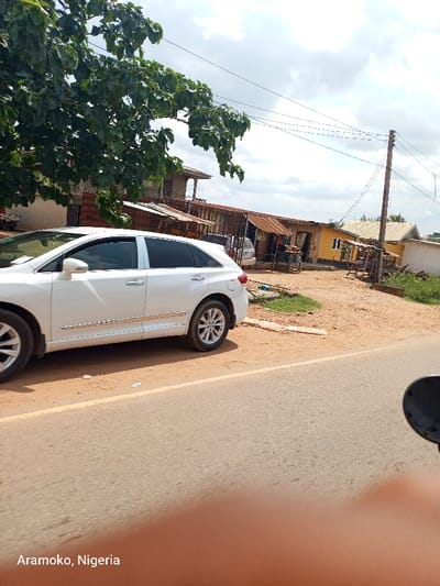 The image shows a white SUV parked on the side of a dirt road in Aramoko, Nigeria. The road is lined with a yellow painted line on the right. The car is facing the direction of the road. In the background, there are several buildings, with some of them being partially obscured by trees. The buildings appear to be residential or commercial. There are power lines above the street. The sky is partly cloudy.
