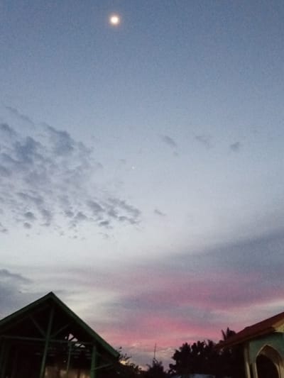The image captures a twilight sky with a bright full moon positioned high in the center. The sky transitions from a pale blue near the moon to hues of pink and purple closer to the horizon, indicating the sun has recently set. Wispy clouds are scattered across the sky, some appearing darker and more defined, while others are lighter and more ethereal.
In the foreground, the silhouettes of two buildings are visible. On the left, the angular structure of a building with a triangular roof and exposed beams is outlined against the colorful sky. To the right, another building with a curved, tiled roof and arched entryway is partially visible. A line of dark trees separates the two structures.
The overall scene suggests a peaceful evening in a tropical or subtropical location, possibly Meuse, Indonesia, given the context. The soft lighting and pastel colors of the sky evoke a sense of calm and tranquility. No text is visible in the image. The activity taking place is the natural progression of day into night.