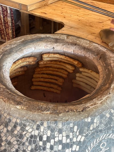 The image shows a tandoor oven with Armenian bread, likely in Tsovagyugh, Armenia. Long loaves are baked vertically against the interior walls of the oven. The exterior of the oven appears to be constructed of stone and is located indoors. Adjacent to the oven is wooden structure. A person's foot is visible at the top left. To the right is a mosaic stone pattern. A rug is visible below and to the left of the wooden structure.