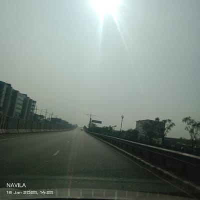 The image captures a perspective from inside a vehicle, looking straight down an elevated highway or expressway in Solepur, Bangladesh. The multi-lane road, paved with dark asphalt, stretches into the hazy distance, marked by faded white lines. Concrete barriers or guardrails line both sides of the thoroughfare.
On the left side of the road, behind the barrier, stand several modern, multi-story buildings featuring glass exteriors, suggesting a commercial or urban development. To the right, dense vegetation, including trees and shrubs, borders the road. Further along the right, a small, isolated single-story building with a light-colored facade is visible amidst the greenery. Utility poles and a large overhead road sign structure, whose text is unreadable, are also present on the right. A faint silhouette of another vehicle can be seen ahead in the distance.
The sky is bright and appears hazy or slightly smoggy, contributing to a muted overall color palette. The sun is prominently positioned in the upper right quadrant, emitting strong, bright rays that create significant glare and diffuse the upper atmosphere.
Visible text overlays in the bottom left corner include "NAVILA" and a timestamp "16 Jan 2025, 14:25," indicating the early afternoon. The scene is devoid of visible people, focusing instead on the infrastructure and atmospheric conditions of the highway.