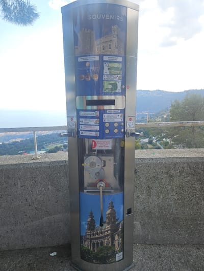 A tall, slender, metallic souvenir vending machine stands outdoors on a textured concrete surface, situated on a high vantage point in Beausoleil, France. The machine's upper section displays "SOUVENIRS" above an image of a stone castle or fortress. It offers two primary functions: a "MEMOEURO® SOUVENIR NOTE" (0 Euro banknote) for 3€ on the right side, and "MEMODAILLE®" (pressed pennies) for 2€ or 1€ plus a customer-provided coin on the left. Instructions for both options are provided in French and English, detailing coin insertion, selection via push-buttons, and a 5-second waiting period after payment. The lower section features a transparent panel revealing internal gears and a silver crank handle with a black grip for the coin pressing mechanism, set against an image of the Monte Carlo Casino building.
The machine is positioned next to a knee-high, rough concrete barrier, topped by a horizontal grey metal railing. Beyond this barrier, a panoramic view unfolds under a partly cloudy sky. To the left, the Mediterranean Sea abuts a densely developed coastal urban area, indicative of Monaco, which includes numerous buildings and sports courts. To the right, tree-covered hills rise in the distance, featuring scattered structures and visible construction cranes. A portion of a dark green pine branch extends into the upper left of the frame. No individuals are present in the scene.
