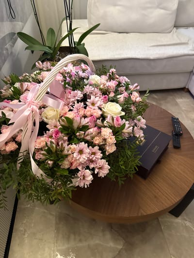 A close-up, eye-level shot focuses on a large basket of pink and white flowers, accented with pink ribbon, resting on a circular wooden coffee table. To the right of the floral arrangement, a dark blue box with gold lettering, possibly a gift box, and two black remote controls are also present on the table. The background reveals a blurred view of a cream-colored sofa and an indoor plant with long green leaves in a pot. The flooring appears to be a light-colored marble-patterned tile. The scene suggests an indoor setting, likely a living room or reception area. The location is specified as Военнослужащих Кп, Russia.