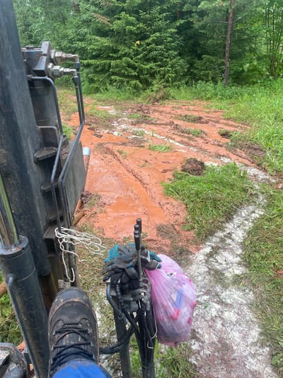 The image depicts a muddy, outdoor scene with a heavy-duty machine in the foreground. The machine appears to be a piece of construction or drilling equipment, with visible hydraulic components, hoses, and a metal arm. A person's black boot, covered in mud, is placed on one of the machine's parts, suggesting an operator is present. They are wearing blue work pants. On the machine, a pink plastic bag filled with what seems to be trash is tied to a piece of equipment. Beside the bag, a pair of dark gloves with blue accents are resting.
The ground is heavily marked with tire tracks, indicating recent activity by heavy vehicles. The mud is a reddish-brown color, suggesting iron-rich soil. Patches of white material are scattered across the ground, possibly construction debris or a substance used in the work. The background features lush green foliage, including coniferous trees and grass, indicating a natural or semi-natural environment.
The overall impression is one of ongoing work in a challenging, muddy terrain, possibly in a rural or forested area. The weather appears to be overcast or the lighting is diffused, as there are no strong shadows. The scene is static, with no clear indication of time of day, other than it is daytime. The presence of the trash bag suggests some form of site work or cleanup. No visible text is present in the image.
