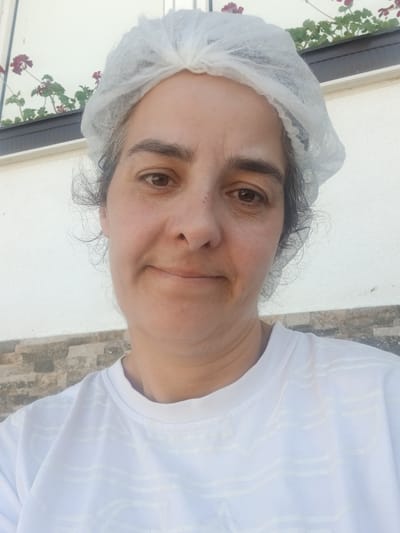 A woman, wearing a white disposable hair net and a white t-shirt, faces the camera. Her hair is dark and curly, peeking out from under the hair net. She has brown eyes and a neutral expression. Behind her, a white wall is visible, with a strip of textured grey and brown tiles running along the bottom. Above the tiles, there is a horizontal dark planter box containing green plants with small red flowers. The scene appears to be outdoors.