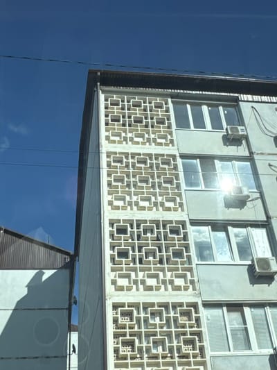 The media file captures a close-up view of a building facade, likely in an urban setting. The building features a distinctive decorative screen composed of repeating geometric square patterns, made of a light-colored material. Above this screen, there are windows with multiple panes, some of which appear to be open. Above the windows, there are air conditioning units mounted on the exterior wall. Wires are also visible running along the top of the building.
The lower part of the image shows a reflective surface, possibly another building or glass, mirroring the sky. The sky is a clear, deep blue, suggesting a sunny day. The lighting indicates it's daytime, with sunlight reflecting off the building's surfaces and the windows. The angle of the shot, looking upwards at the building, and the reflective surfaces suggest the photo might have been taken from a lower vantage point, possibly from across a street or from a different building. The architectural style, particularly the decorative screen, might offer clues to the era or region the building is from. No people or specific activity are evident in the frame. The overall impression is one of architectural detail and the interaction of the building with its environment.